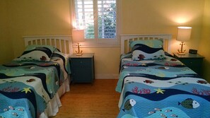 A room with two beds, each with a blue ocean-themed bedspread, a small nightstand between them, and a window with blinds. - Charming Remodeled Single Level. HotTub. Fenced backyard. Porch view! (Cambria)