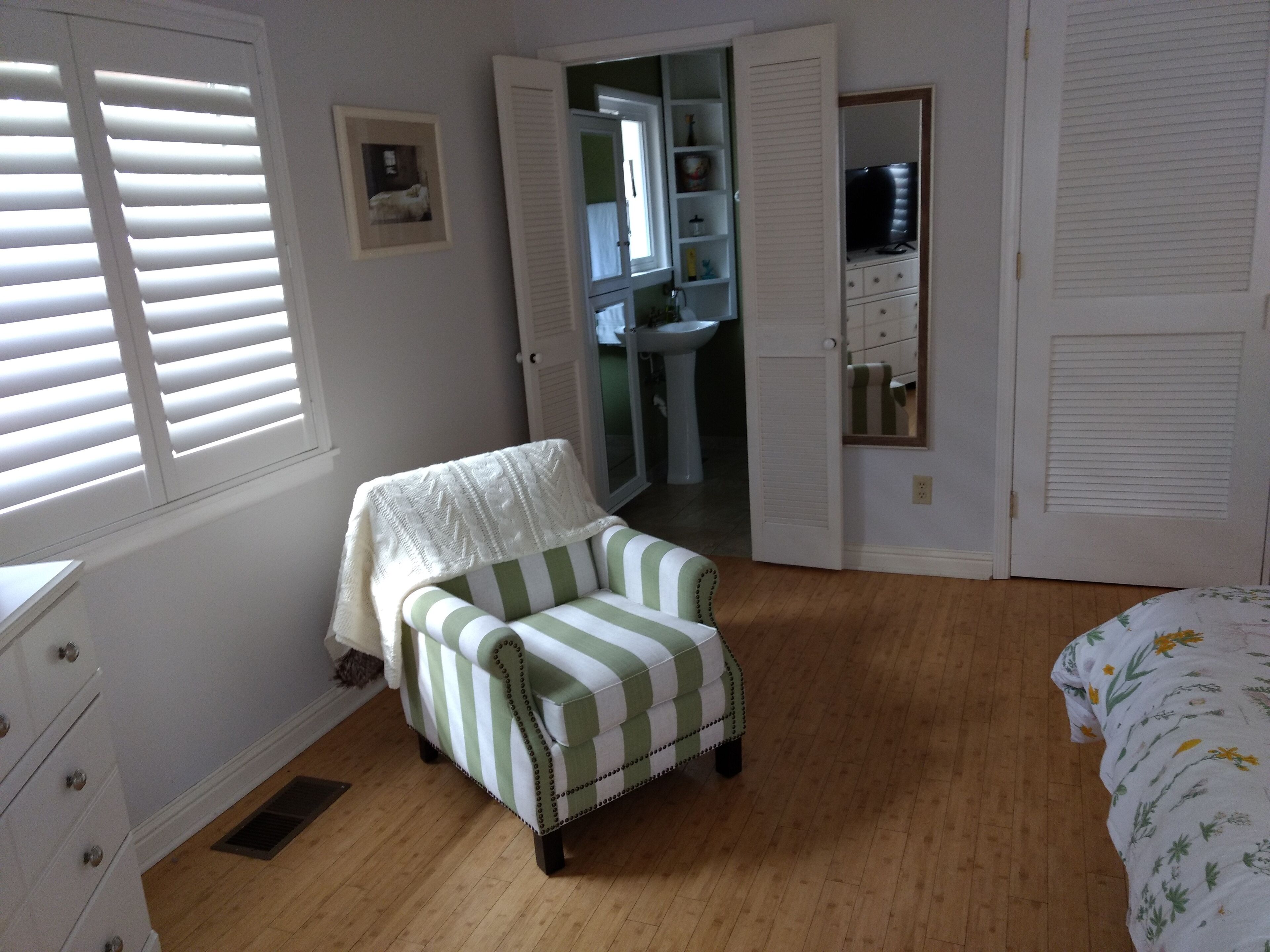 Een slaapkamer met een gestreept fauteuil, een bed met bloemenbeddengoed, een kast en een raam met jaloezieën.