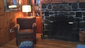 Ein gemütliches Wohnzimmer mit Holzwänden, einem Kamin aus Stein, einem Bücherregal und einem bequemen Sessel.