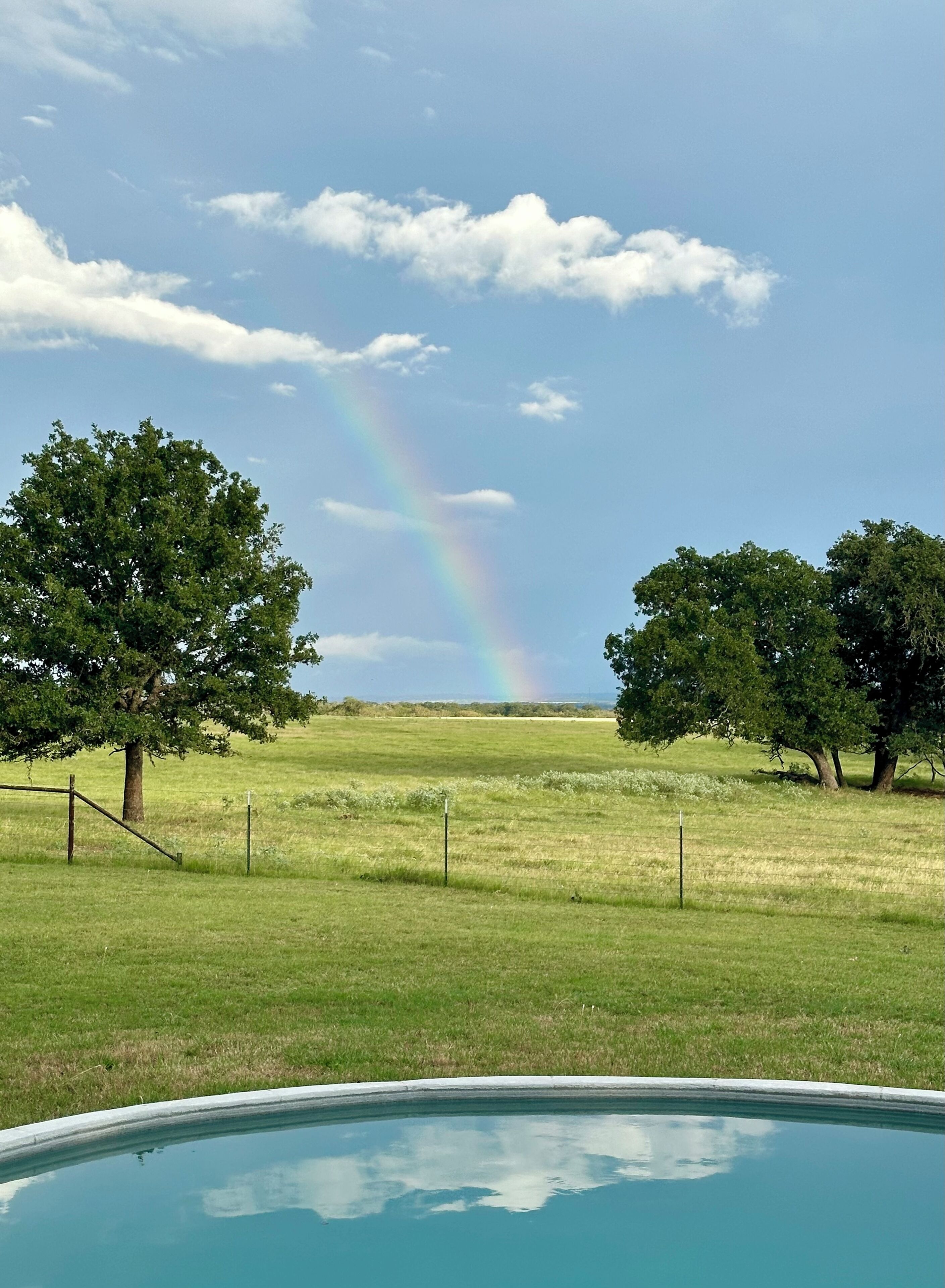 A grassy field with a rainbow, trees, and a swimming pool.