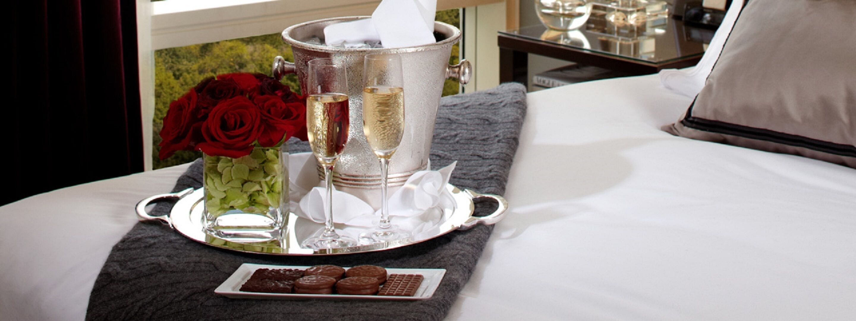 Un plateau avec des coupes à champagne, un vase de roses rouges et une assiette de biscuits posée sur un lit.