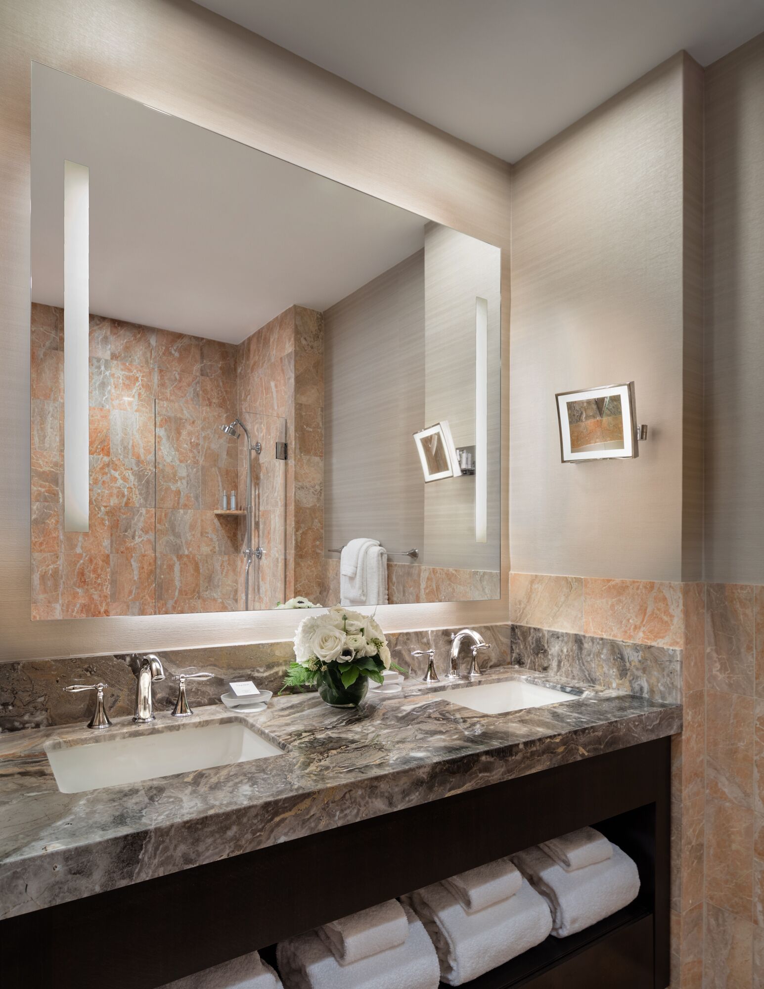 A modern bathroom with a large mirror, two sinks, marble countertops, and a towel rack with towels.
