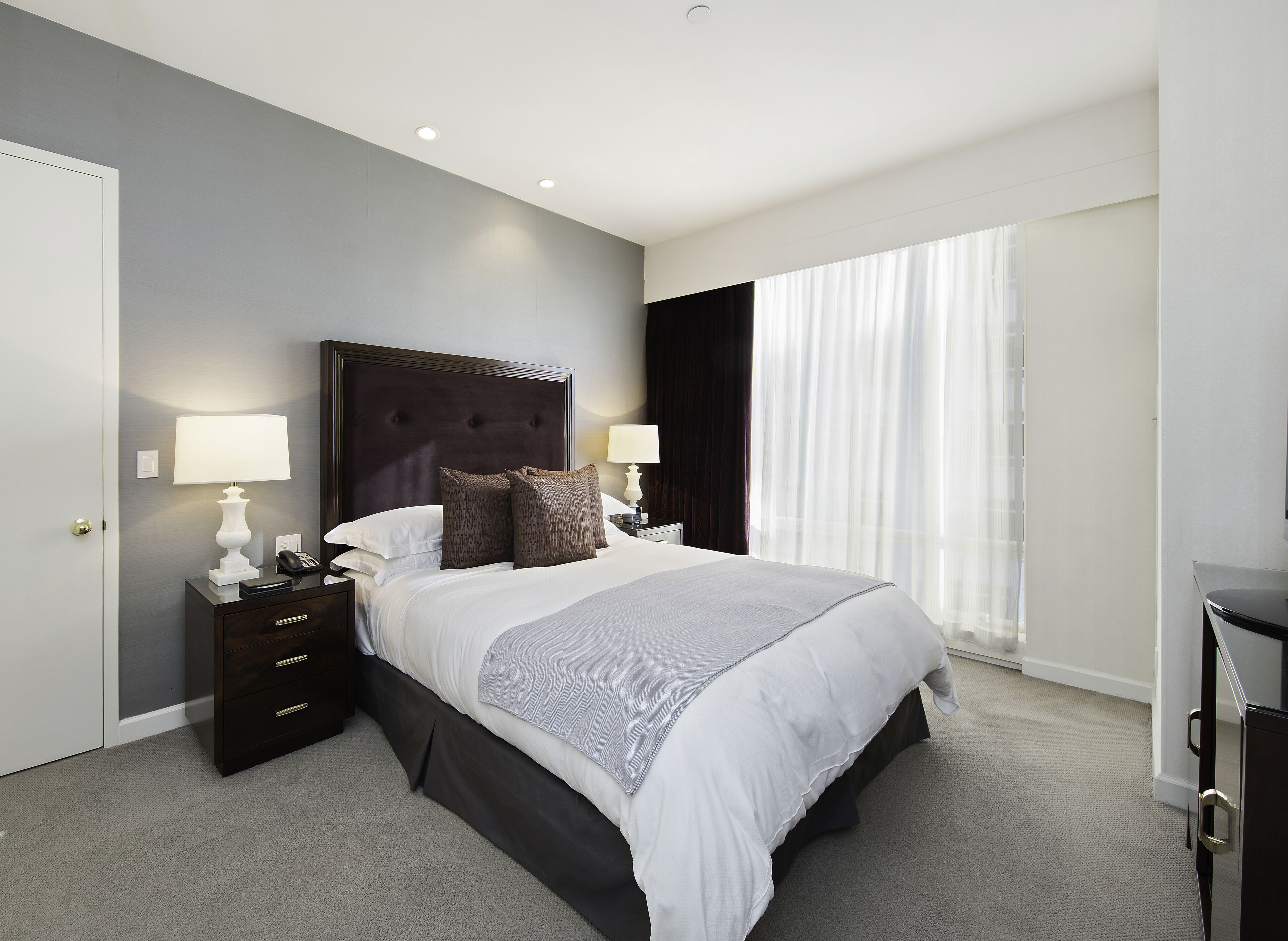 Una habitación de hotel moderna con una cama grande, dos mesitas de noche con lámparas, un cabecero oscuro y un ventanal con cortinas translúcidas.