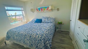 Ein Schlafzimmer mit Bett, Fenster, Wandlampe, einem kleinen Tisch und einer Kommode.