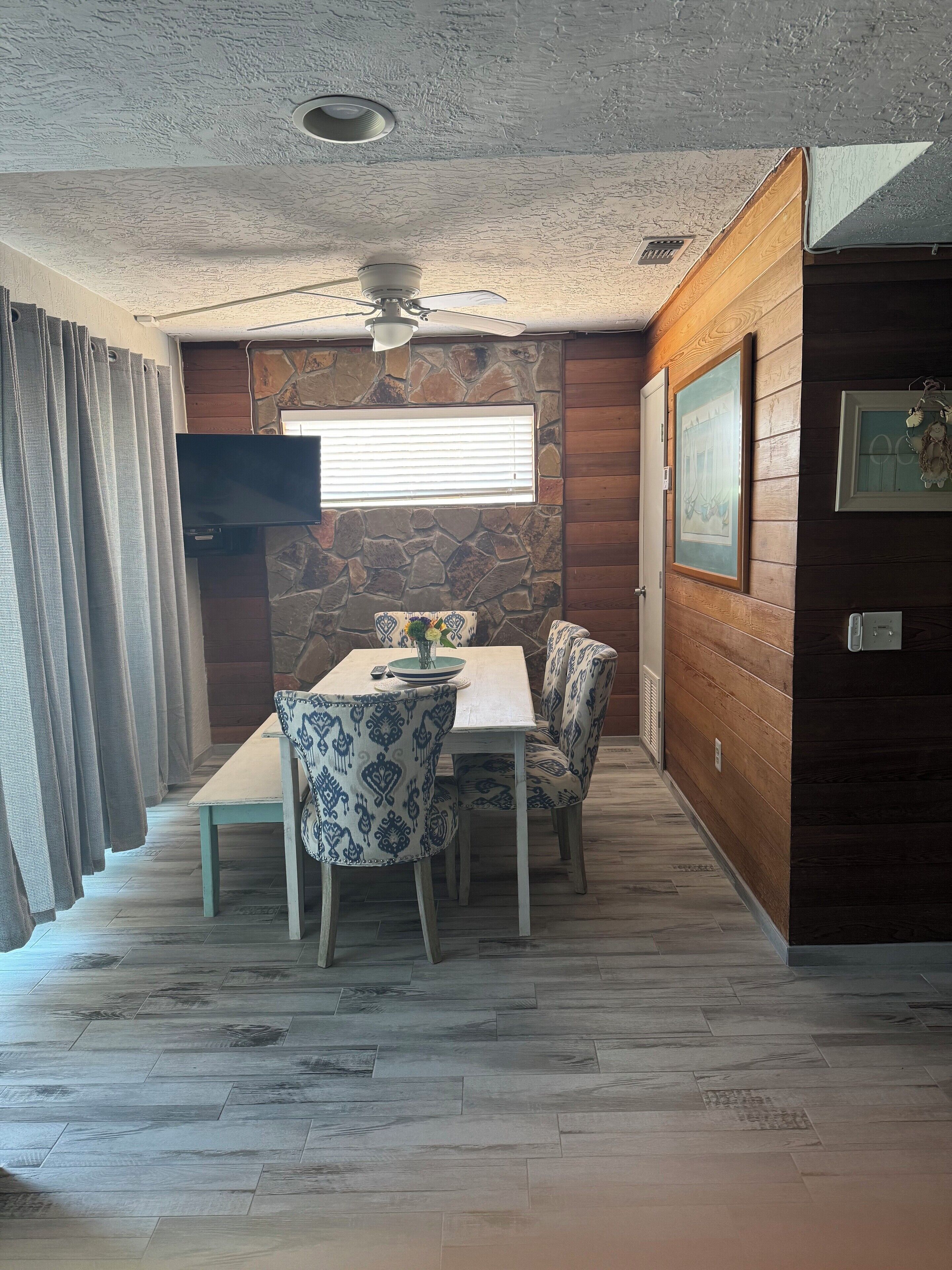 Un comedor con una mesa y sillas, un ventilador de techo, una pared de piedra y una ventana con persianas.