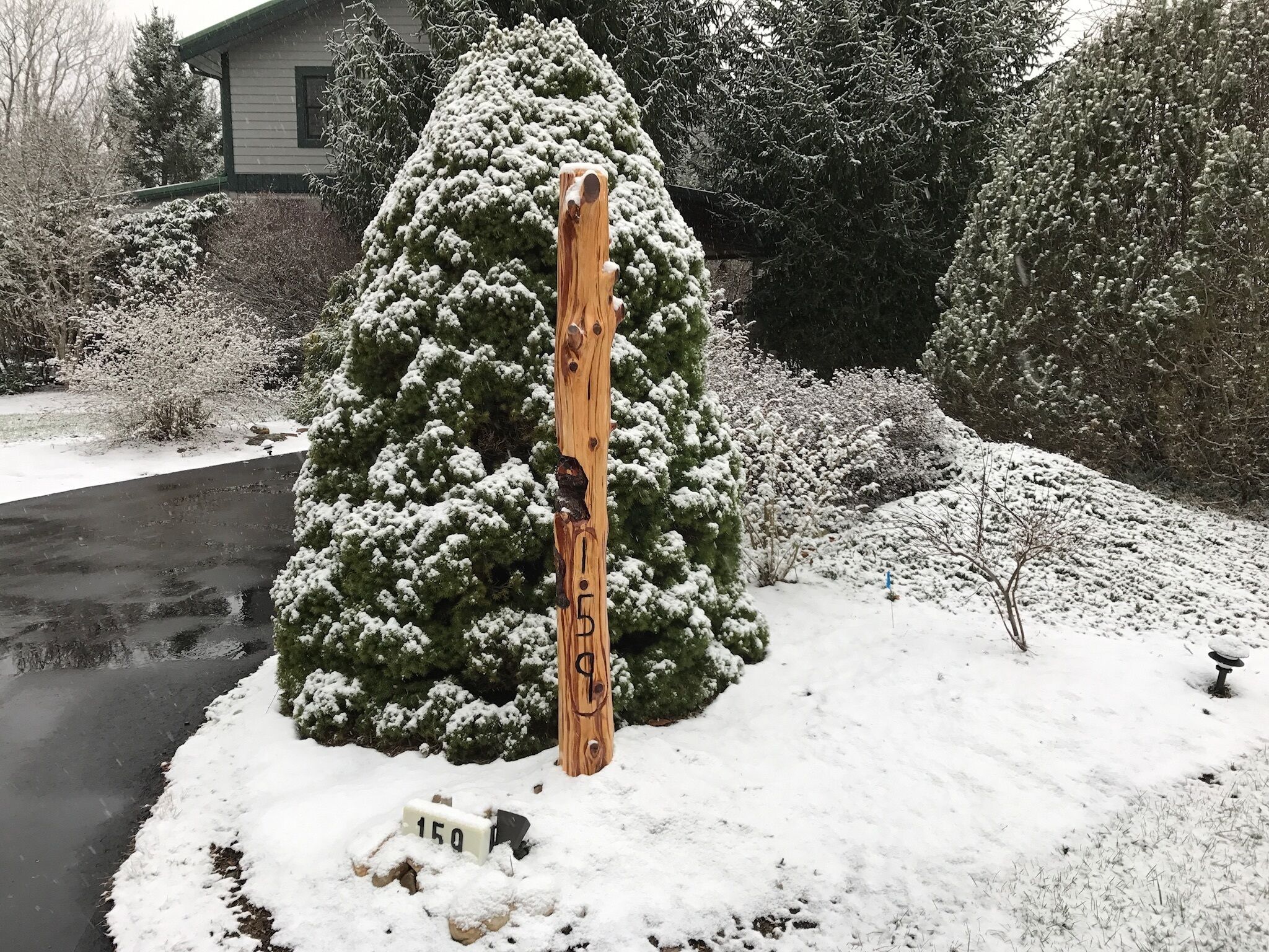 A wooden post with a carved figure, a snow-covered evergreen tree, and a house in the background.