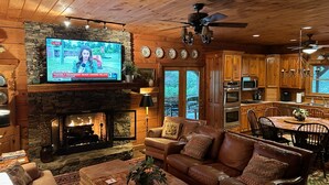A cozy living room with a stone fireplace, leather sofa, and a television displaying a news broadcast. - Beautiful Log Cabin with river access to The Watagua. Newly  added Fire Pit (Banner Elk)