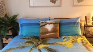 A bed with a tropical beach-themed bedspread, a potted plant, and framed artwork on the wall. - BOOK 30 DAYS OCT 15-NOV FREE PARKING! UNIT 52 WALK TO INTL MKT, TARGET, BEACH (Honolulu)
