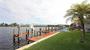 Un muelle se adentra en aguas tranquilas, rodeado de un césped cuidado y una palmera.