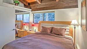 Un dormitorio con techo de madera, una cama con cabecera, lámparas de noche, una mesita de noche, un espejo y una ventana con vistas a árboles.