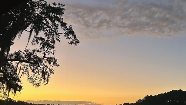 A glass of white wine on a balcony overlooking a sunset over a water body with trees and a distant horizon.