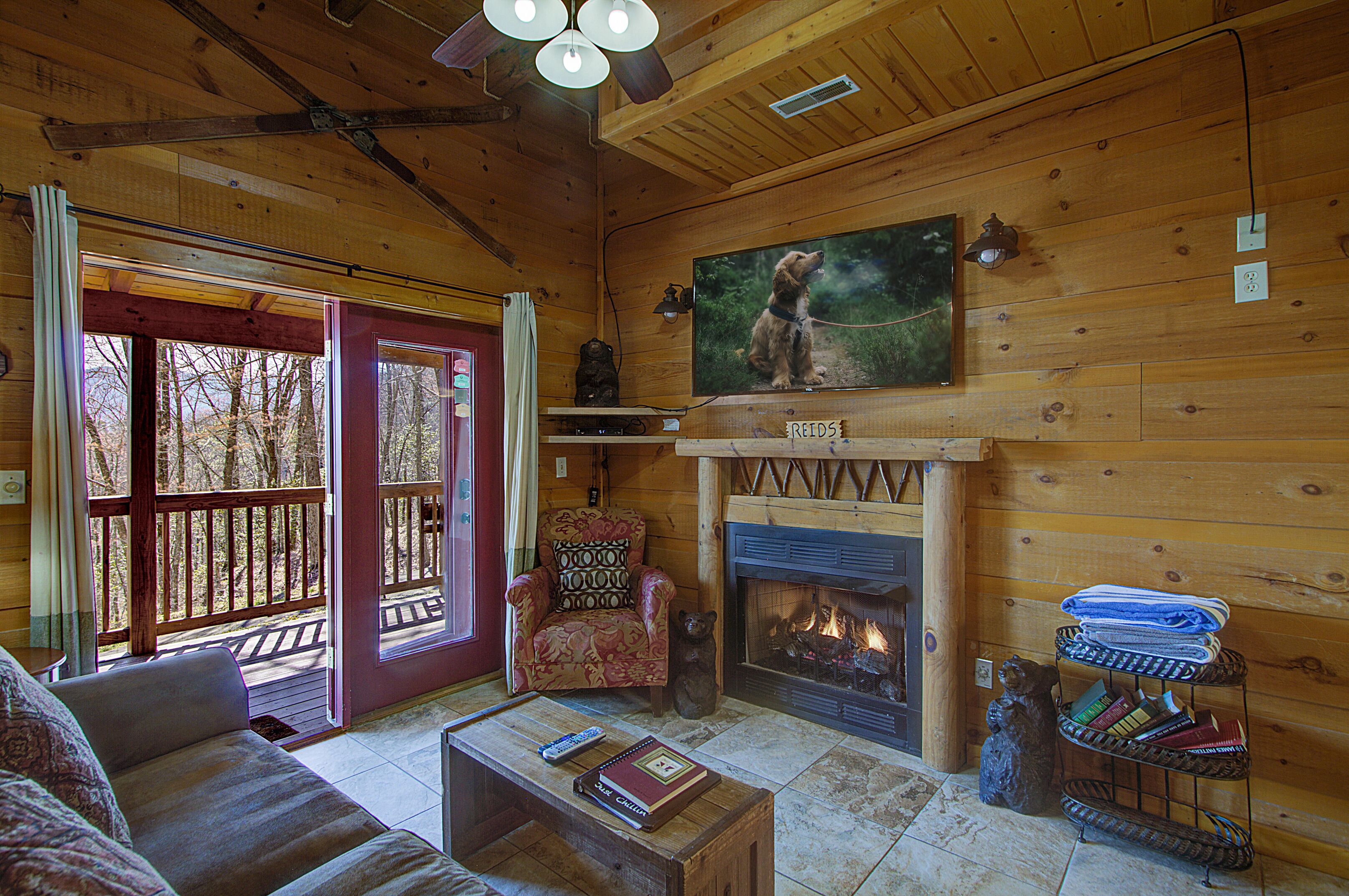 A cozy cabin interior with a fireplace, a television mounted above it, a comfortable sofa, and a wooden deck visible through the glass door.
