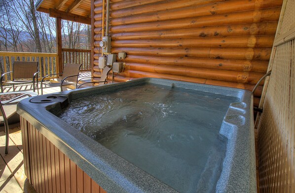 Un jacuzzi en una terraza de madera con sillas y una cabaña de madera al fondo.