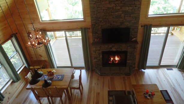 A cozy living room with a stone fireplace, wooden floors, and large windows offering views of the outdoors.