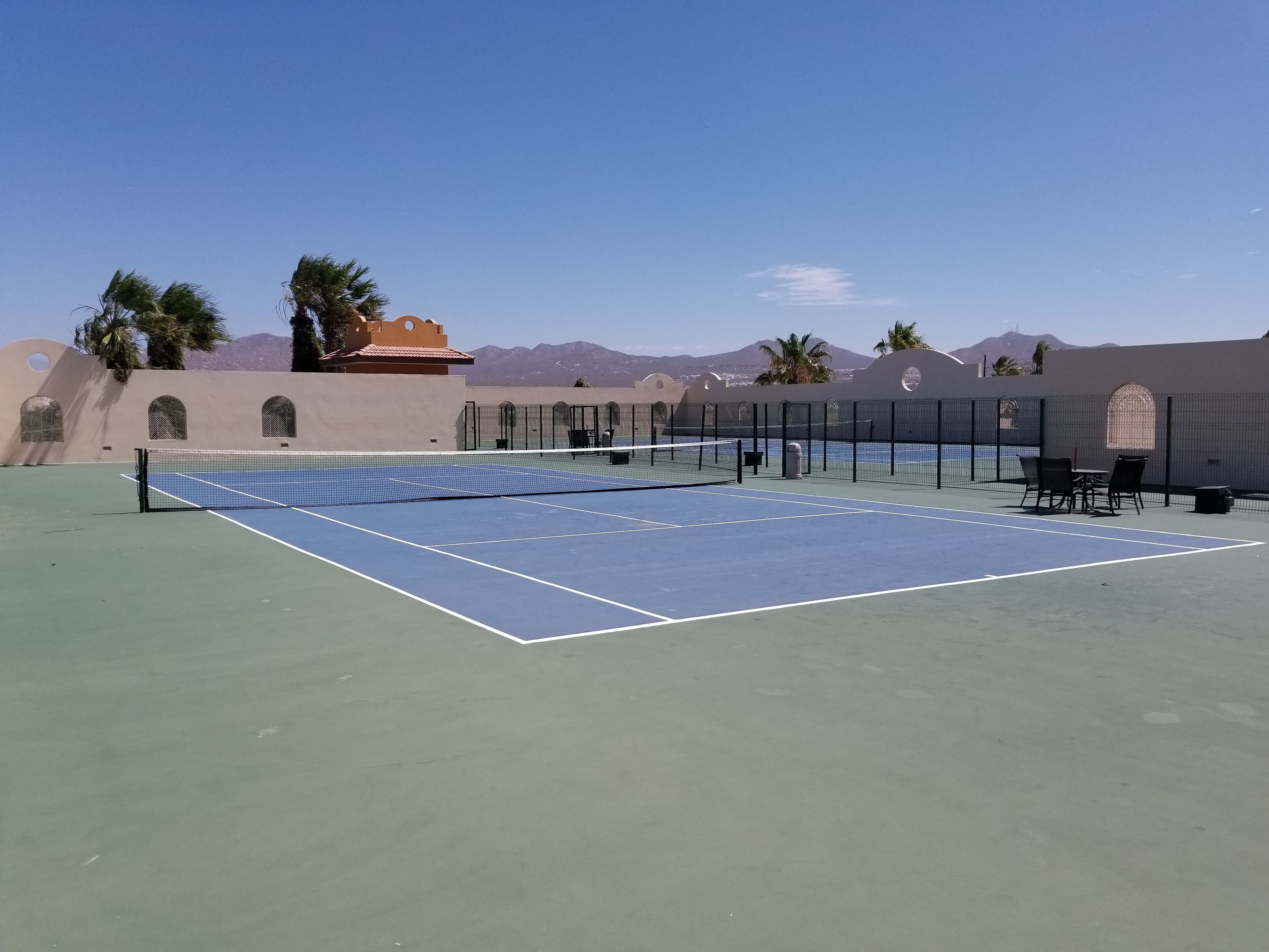 A tennis court with a net, surrounded by a fence, and a seating area with chairs.