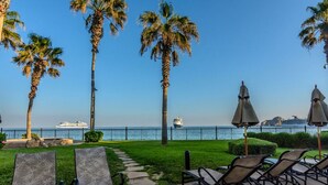Un espace patio aménagé avec des chaises longues et des parasols, des palmiers, et une vue sur la mer, avec un navire de croisière visible au loin.