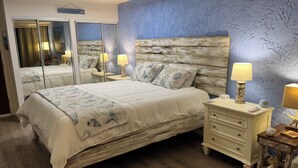 Un dormitorio con una cama grande, cabecera de madera, dos mesitas de noche con lámparas, un espejo y una pared azul con textura.