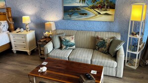 Una habitación con un sofá a rayas, una mesa de centro de madera, una mesita de noche con una lámpara y un cuadro de una playa en la pared.
