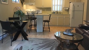 A compact kitchen with white cabinets, a microwave, and a refrigerator. There is a dining area with a glass table and black chairs. A decorative statue is on the glass table. - Bayfield- Charming Condo, Jumping Off Point to Apostles (Bayfield)
