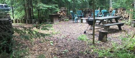 A forested area with picnic tables and chairs, a barbecue grill, and a stack of firewood.
