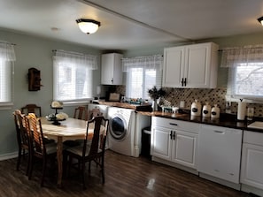 A kitchen with white cabinets, a wooden dining table with chairs, a washing machine, and a countertop with kitchen appliances.