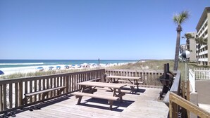 A wooden deck with picnic tables overlooking a beach with umbrellas and a clear blue sky. - Emerald Isle 107-On the Beach-Ground Floor (Fort Walton Beach)