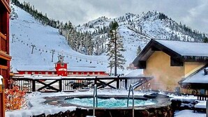 Escena nevada de una montaña con un telesilla, una zona de piscina y un edificio de techo rojo.