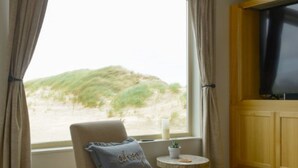 Una habitación con un ventanal grande, una silla con almohada, una mesita con vela y un televisor sobre un mueble de madera.