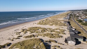 Zona costera con playa, dunas y una hilera de casas a lo largo de una carretera.