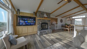 Amplia sala de estar con chimenea de piedra, vigas de madera, un televisor grande y comedor con vistas.