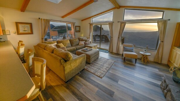 Amplia sala de estar con un sofá grande, una mesa de centro y un comedor con mesa y sillas. Amplias ventanas ofrecen vistas al exterior.