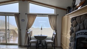 Una habitación con un ventanal grande que da a una playa, una chimenea de piedra y una mesa con sillas.