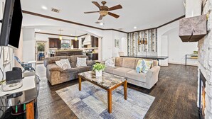 A spacious living room with a large sofa, a coffee table, and a TV. There is a kitchen in the background with cabinets and a dining area. - Fam Friendly 4 BDRM Home - Walk to Shops & Bars I Mins To DT & Convention Center (Dallas)
