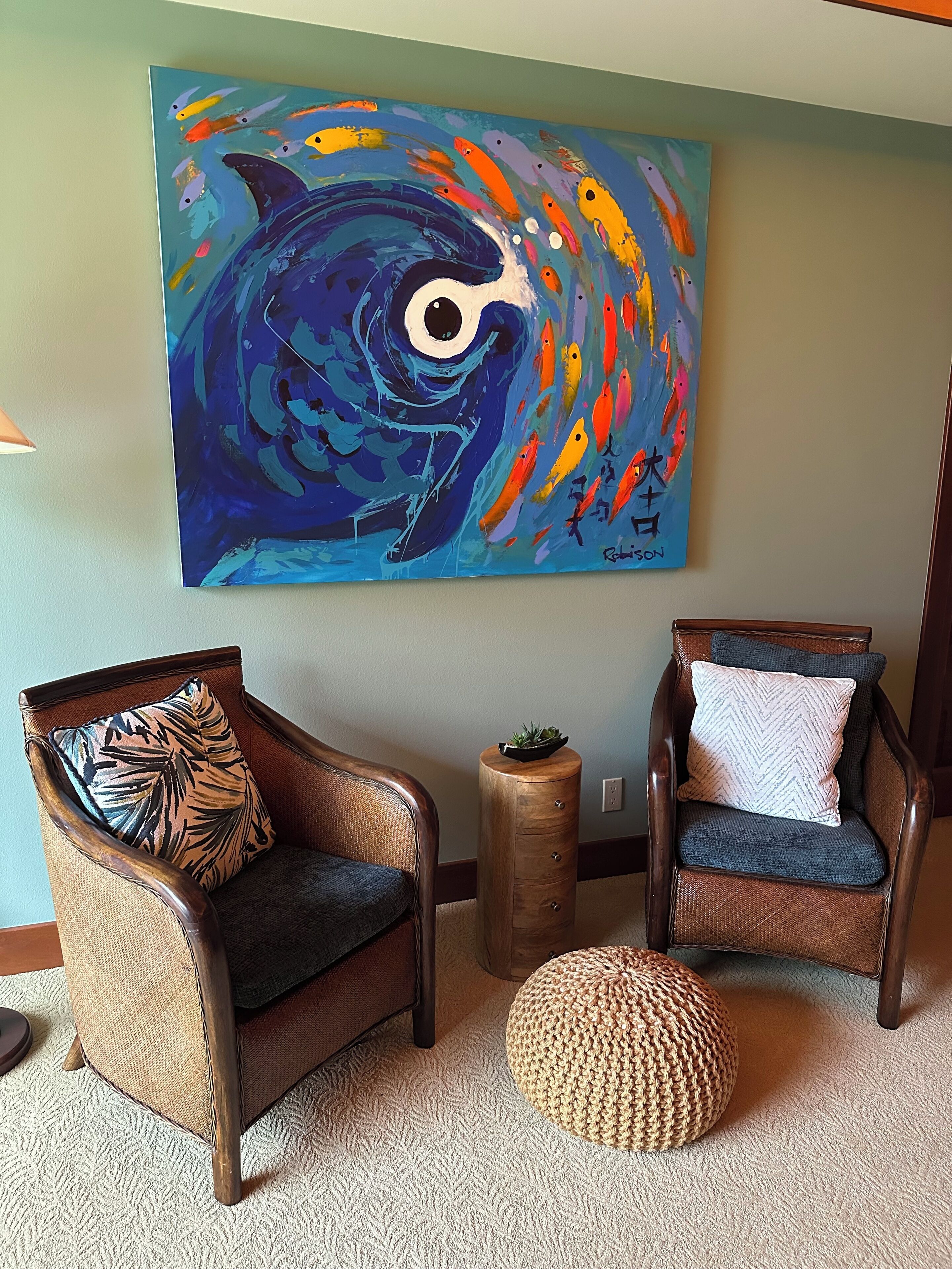 A room with two wicker chairs, one with a leaf-patterned pillow and the other with a chevron-patterned pillow. A round woven ottoman is on the floor. A large abstract painting with fish and a whale is on the wall.