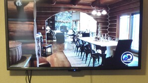 A flat-screen TV mounted on a wall displaying a dining area with a long table and chairs, a stone wall, and a wooden ceiling. Below the TV, there is a digital clock and a shelf with books.
