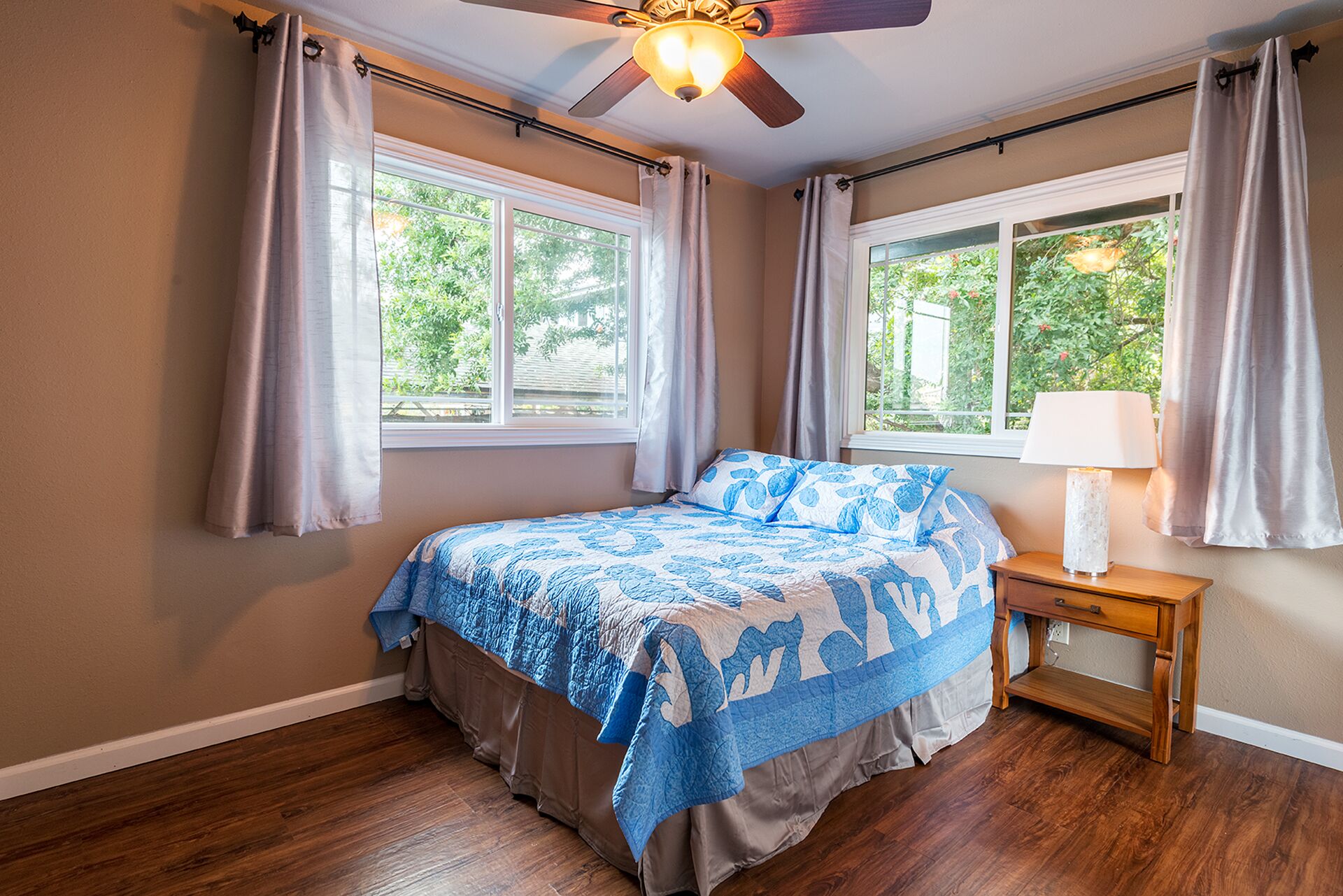 Quarto com cama, duas janelas, ventoinha no teto e mesa de cabeceira com abajur.