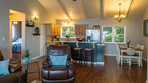 Un vaste salon doté d’un canapé en cuir, un coin repas avec une table et des chaises blanches, une cuisine avec des meubles en bois et un réfrigérateur, ainsi qu’un ventilateur de plafond.