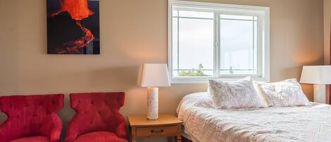 Een slaapkamer met een bed, twee rode stoelen, een nachtkastje met een lamp, een raam en een schilderij van een vulkaan.