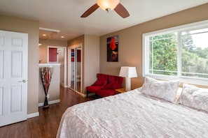 A bedroom with a large bed, a red chair, a ceiling fan, a window with a view of trees, and a bathroom visible through an open door.