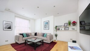 A modern living room with a beige sectional sofa, a red shag rug, and a white coffee table. There are framed pictures on the walls and a small plant on a shelf. - 110 m² luxury two-bed apartment in Earls Court (London)