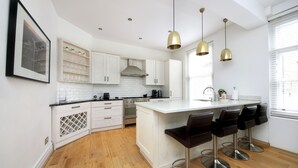 A modern kitchen with white cabinetry, a central island with bar stools, pendant lighting, and a large window providing natural light. - 110 m² luxury two-bed apartment in Earls Court (London)