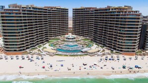 A high-rise hotel with multiple balconies, a central pool area, and a beach with sun loungers.