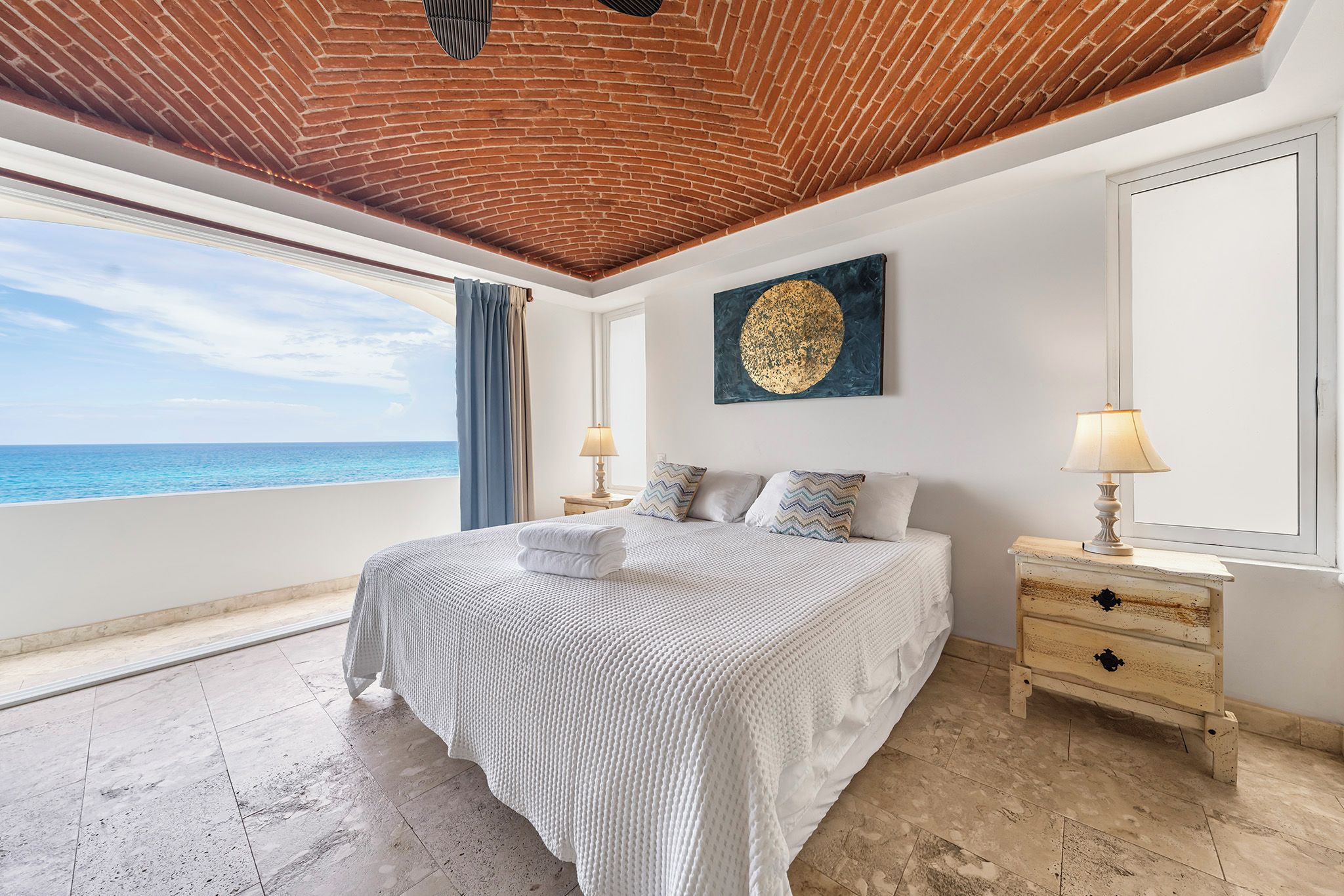 Ein Schlafzimmer mit einem großen Bett, einem Nachttisch, einer Lampe, einem Gemälde und Blick auf das Meer.