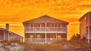 Une maison de deux étages située en bord de mer, dotée d’un balcon et entourée de sable et de végétation.