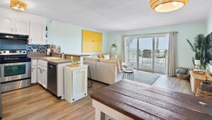Une cuisine moderne avec des armoires blanches, un four en acier inoxydable et un lave-vaisselle. On y trouve un coin repas avec une table en bois et un banc. Une grande fenêtre offre une vue sur l’extérieur.