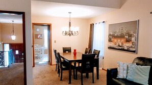 A dining area with a chandelier, a dining table with chairs, a black leather sofa, and a cityscape painting on the wall.