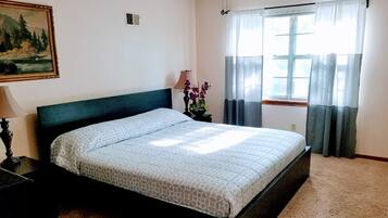 Een slaapkamer met een bed, nachtkastje, lamp, schilderij, raam met gordijnen en een plafondventilator.
