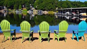 Una hilera de sillas Adirondack verdes y azules mirando a un lago con un muelle y barcos.