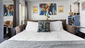 A modern hotel room with a large bed, a desk, and a chair. There are abstract paintings on the wall and a guitar on the desk.