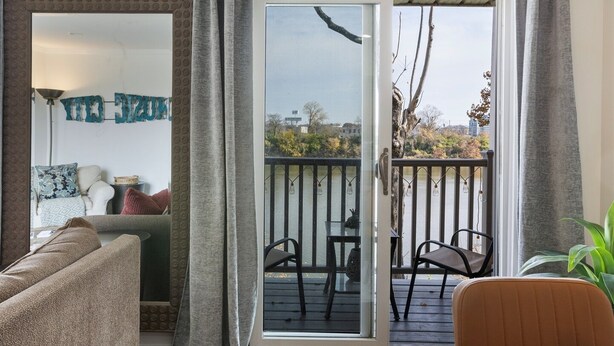 A living room with a sofa, a coffee table, and a dining table. There is a sliding glass door leading to an outdoor balcony with a railing.